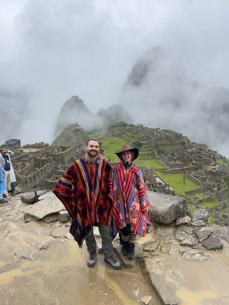 Dos viajeros con ponchos andinos de colores posan frente a Machu Picchu entre la neblina