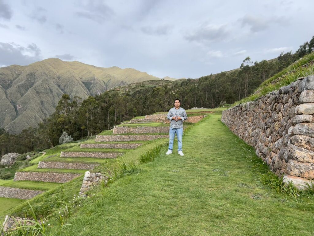 Persona de pie en un sendero de césped junto a andenes incas de piedra, con montañas al fondo, en Cusco