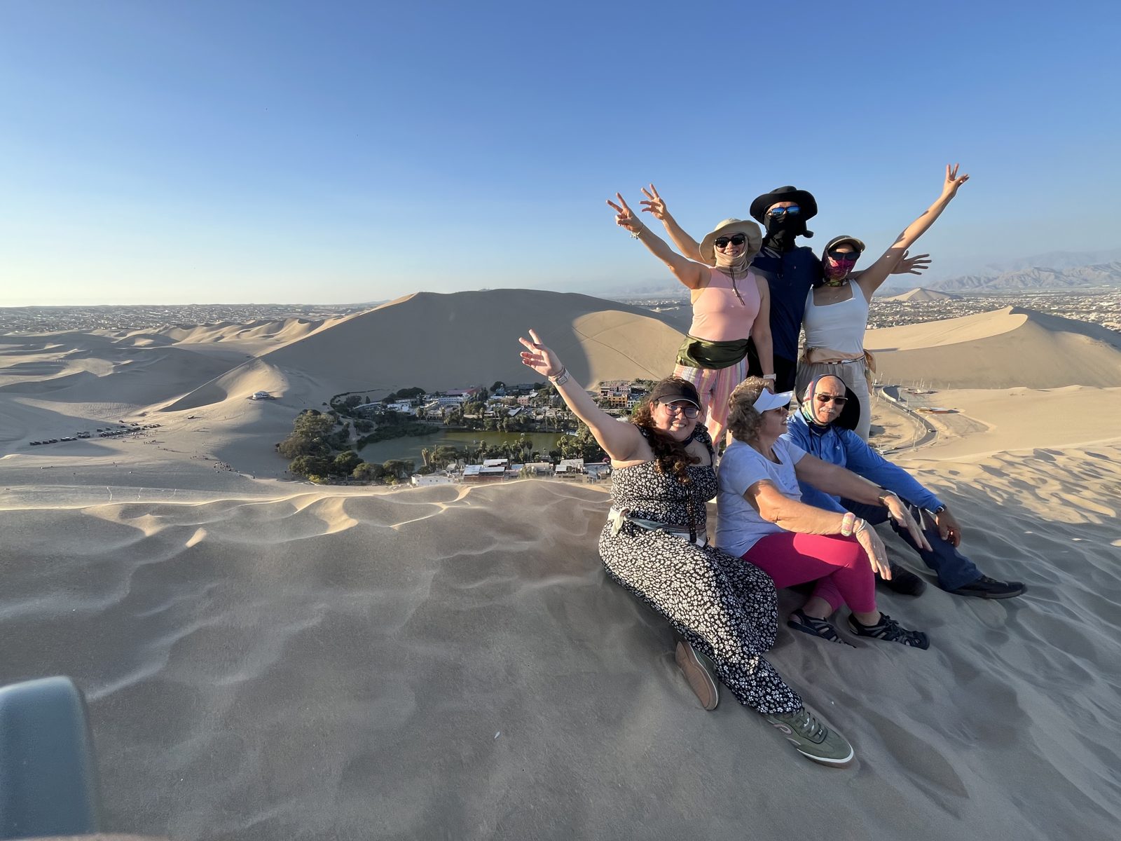 Grupo de viajeros con los brazos en alto sobre una duna frente al oasis de Huacachina